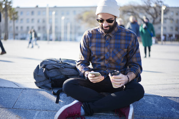 Young bearded male traveler resting with coffee to go sitting on urban setting squaire booking hostel via smartphone and 4G internet, handsome travel blogger updating multimedia on site using mobile - Foto, afbeelding