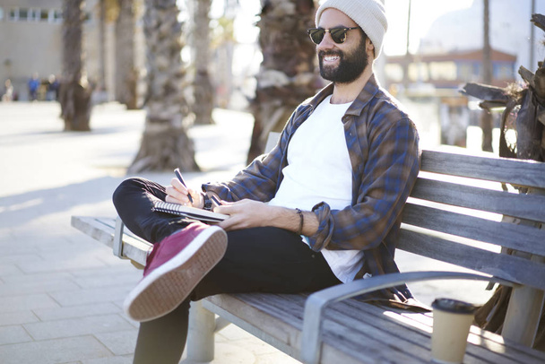 Cheerful bearded guy in sunglasses sitting on bench spending free time on hobby making sketches in notepad, smiling male tourist creating travel diary while resting after city tour enjoying trip - Фото, изображение