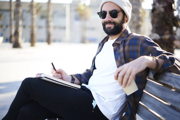Portrait of handsome bearded male travel journalist in sunglasses and casual outfit sitting on bench writing article in notepad satisfied with working process outdoors and spring sunny weather - Фото, зображення