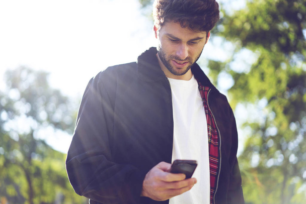 Positive good looking student checking notification while sending messages to friends using application on smartphone connected to internet.Handsome young tourist spending leisure time outdoors - Foto, Imagem