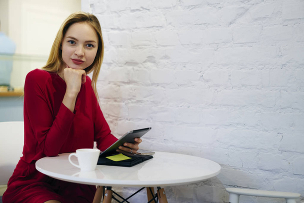 Portrait of attractive blonde female entrepreneur sitting in cafe interior using modern technology for browsing, confident businesswoman looking at camera while checking notifications on touchpad - Fotó, kép