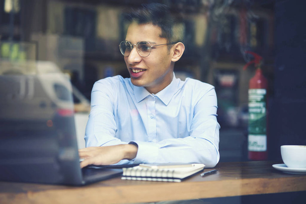 Positive young male student in stylish eyeglasses watching funny video on laptop computer connected to internet.Smiling man reading news in websites and browsing information sitting in cafeteri - Photo, Image