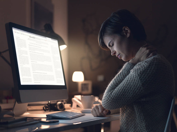 Young woman working with her computer late in the office and suffering from neck pain - 写真・画像