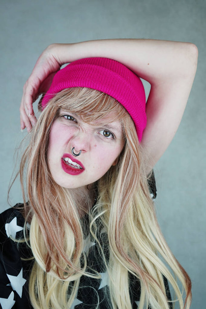 Портрет молодой блондинке с пирсинг в нос в розовой шапке на фоне - Фото, изображение