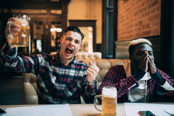 Handsome young man holding his beer and happy shouting looking at the TV screen watching football game with his upset afro american friends at the local pub while support different teams. - Foto, imagen