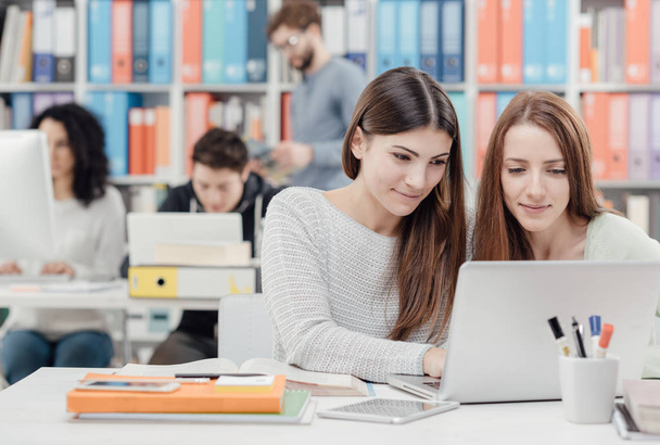 Jóvenes estudiantes en la biblioteca, están estudiando juntas y conectándose con un portátil
 - Foto, Imagen