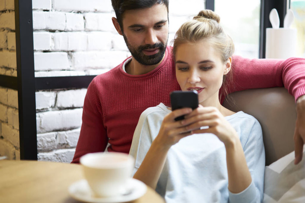 Young good looking couple in love chatting with followers in social networks using application on telephone.Pondering male and female reading news in websites on internet while sitting in cozy place - Photo, Image
