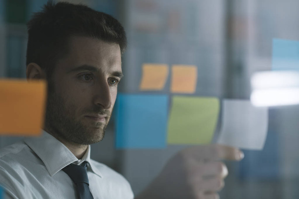 Business executive reading sticky notes on a glass, he is thinking about creative business solutions and strategies - Foto, Imagem