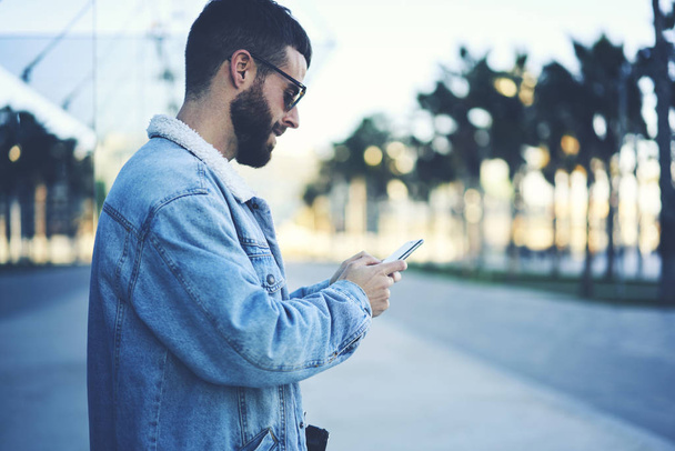 Handsome bearded male in good mood reading news on web pages and texting feedback for web store via modern telephone connected to 4G internet,young blogger walking at free time in urban stree - Photo, image