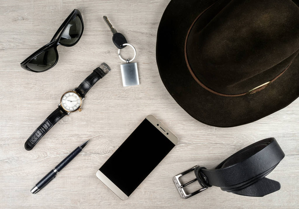 Smartphone, chapéu de feltro, relógio de pulso, cinto de couro e acessórios de outros homens na superfície com uma textura de carvalho branqueado
. - Foto, Imagem