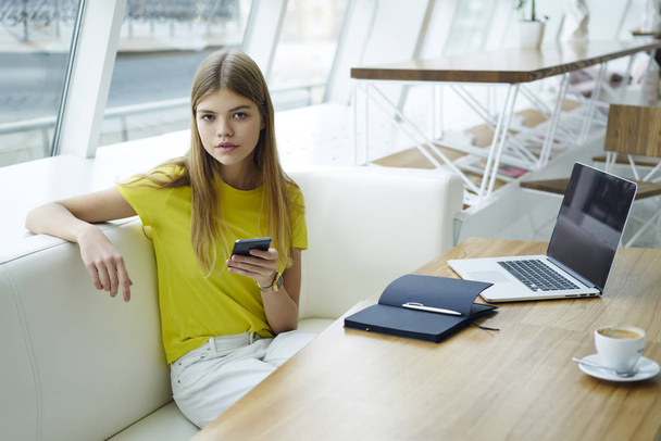 Portrait of beautiful hipster girl dressed in yellow t-shirt holding telephone in hand and looking at camera while sitting on sofa at wooden table with laptop computer and opened notepad.Mock up area - Foto, afbeelding