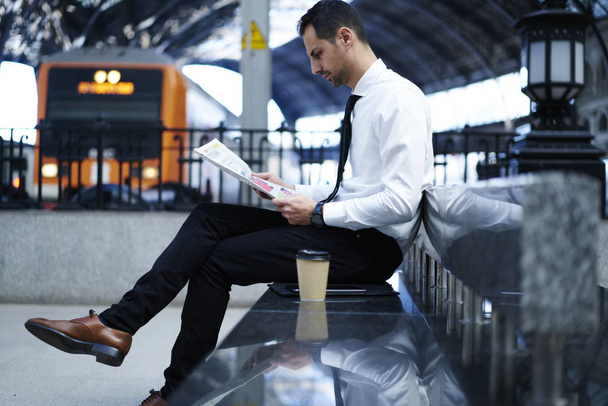 Handsome businessman in elegant formal outfit waiting for train in railway station reading financial news,pensive male manager spending time checking daily newspaper articles while staying on platform - Valokuva, kuva