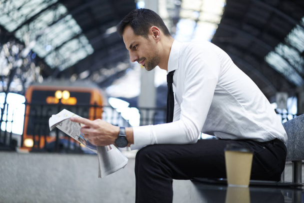 Side view of formally dressed businessman concentrated on reading articles in daily newspaper sitting near platform, prosperous financial manager waiting in railway station for public transport - Фото, изображение