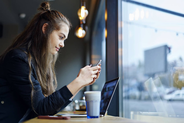 Mladá krásná žena grafik oblečený v odesílání zpětné vazby na mobil připojen k 4g, zatímco sedí u stolu s chutný nápoj v skla a přenosný počítač vnitřní stylové oblečení - Fotografie, Obrázek
