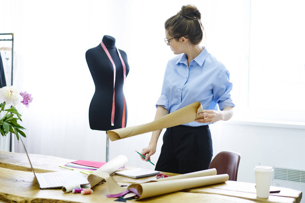 Trendy dressed female dressmaker looking at mannequin creating sketches new collection during working process in showroom, talented designer standing near desktop analyzing measures for blueprint - Fotoğraf, Görsel