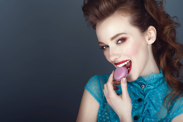 Close up portrait of a beautiful model with creative hairstyle and colourful make up eating violet macaroon and smiling - Foto, Imagen