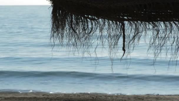Sombrilla de playa de paja con olas del mar rompiendo en la orilla en una mañana soleada
 - Imágenes, Vídeo