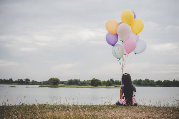 Little cute girl sitting on long green grass outside. Girl holding colorful balloons in hand.  - Foto, imagen