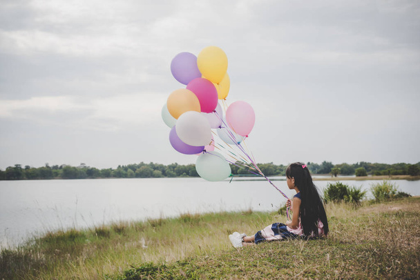 Little cute girl sitting on long green grass outside. Girl holding colorful balloons in hand.  - Photo, image