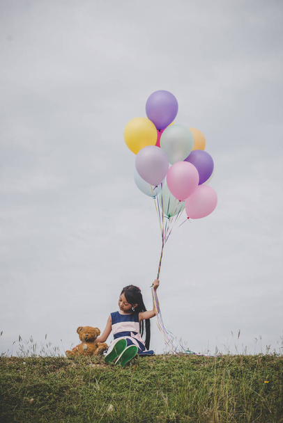 Little cute girl with teddy bear sitting on long green grass outside. Girl holding colorful balloons in hand.  - Fotoğraf, Görsel