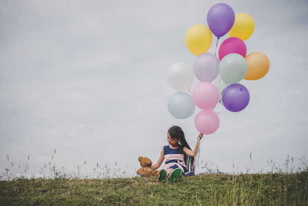 Little cute girl with teddy bear sitting on long green grass outside. Girl holding colorful balloons in hand.  - 写真・画像