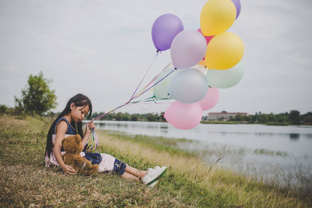 Roztomilá holčička s Medvídek sedící na dlouhé zelené trávě mimo. Dívka v ruce drží barevné balónky.  - Fotografie, Obrázek