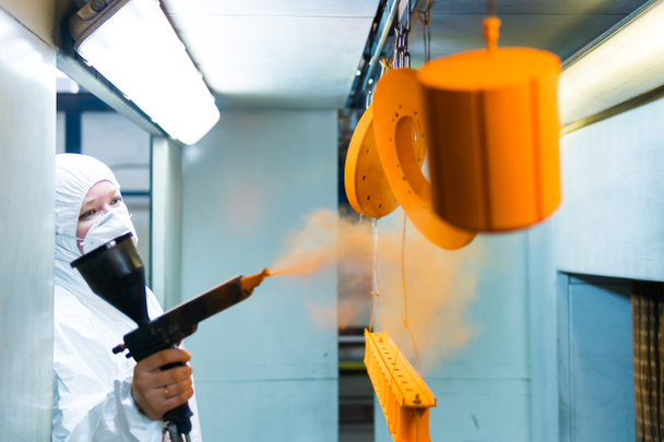 Metalliosien jauhemaalaus. Nainen suojapuvussa suihkuttaa jauhemaalia aseesta metallituotteisiin
 - Valokuva, kuva