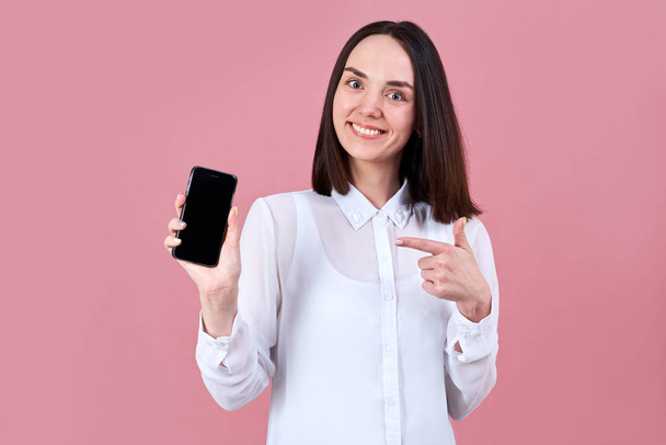 young friendly brunette girl friendly smiles widely, pointing his finger at the smartphone in her hand.  - Fotó, kép