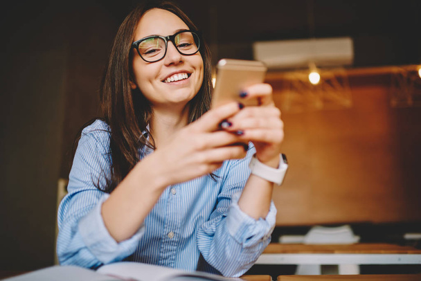 Portrait of cheerful brunette woman checking mail on smartphone getting discount for shopping in web store,happy woman in eyeglasses having fun watching photos  and video on mobile phone in cafe - Zdjęcie, obraz