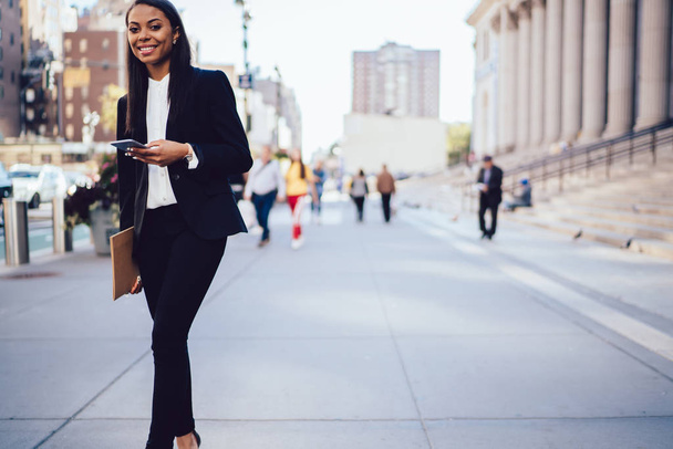 Portrait of successful African American student in black stylish suit smiling at camera while walking outdoors in urban setting with cellphone. Cheerful businesswoman standing near office building  - Photo, Image