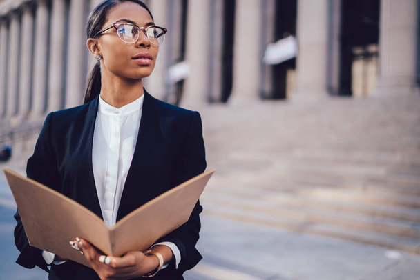 Thoughtful dark skinned female financial manager in black suit holding folder in hands while standing against office building outdoors. African American student of high financial economist university - Foto, Bild