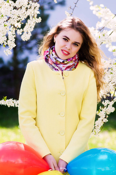 Una joven vestida con un abrigo amarillo de moda en un parque floreciente. Sostiene globos
. - Foto, imagen