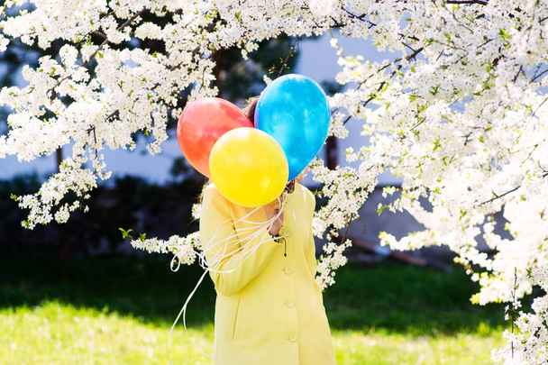 Hava balonları arkasında bir çiçekli parkta gizli bir moda sarı ceket giymiş genç bir kadın - Fotoğraf, Görsel