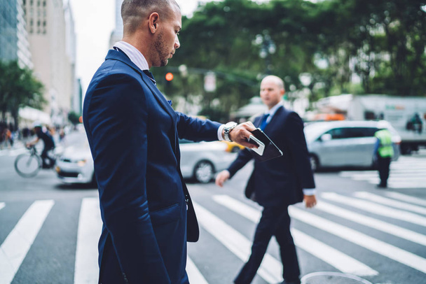 Caucasian male lawyer looking at wristwatch crossing street and getting to office by foot in time, side view of confident businessman checking time planning schedule walking in downtown on crosswalk - 写真・画像