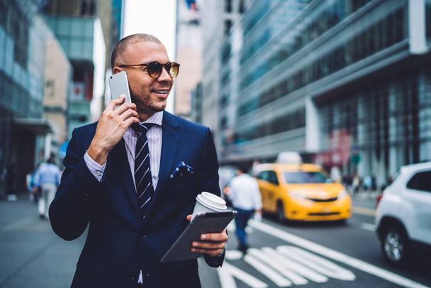 Smiling executive manager in sunglasses talking on phone while strolling with touchpad in business district, prosperous male lawyer having mobile conversation about work holding tablet  checking mail - Фото, изображение