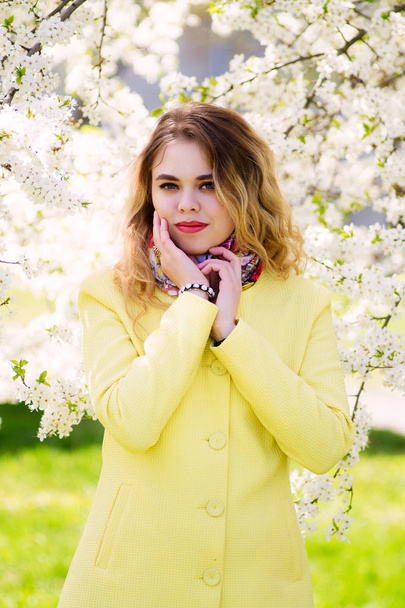 Egy fiatal nő öltözve egy divatos sárga kabátot, egy virágos parkban. nézi a kamera. - Fotó, kép