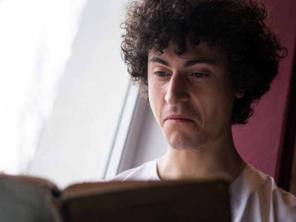 Mann mit lockigem Haar liest vor dem Fenster ein Buch - Foto, Bild