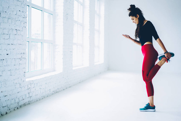 Young woman training in gym watching video from web page online on mobile for stretching muscles of lower body,sportswoman warming up and blogging in networks on smartphone standing near publicity area - Fotografie, Obrázek