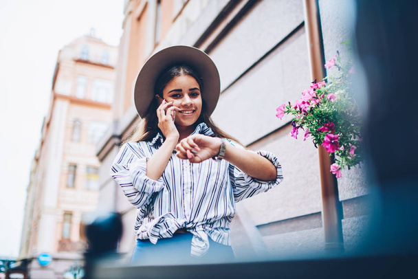 Half length portrait of positive fashionable young woman in trendy hat managing time while satisfied with good news about mobile benefits during telephone conversation strolling on street of city - Foto, afbeelding