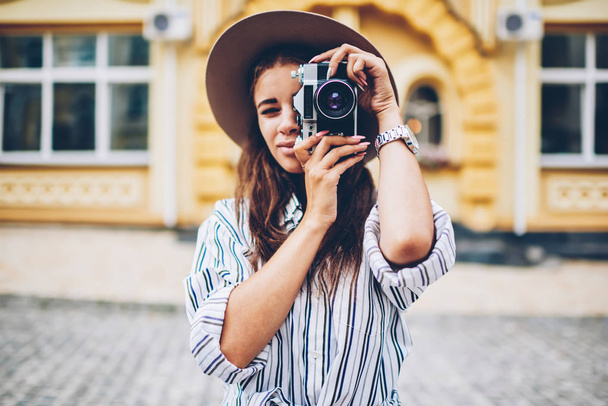 Half length portrait of stylish female tourist in trendy hat focusing and making photo on vintage camera with modern lense standing on architectural promotional background of downtown - Photo, Image