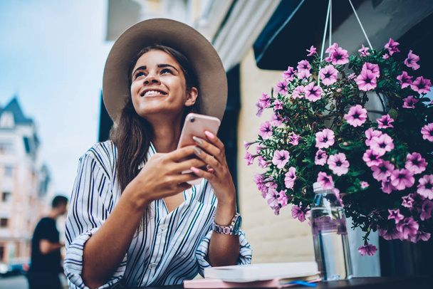 Cheerful dreamy stylish student wearing cool hat and looking up while received notification with good news on smartphone.Positive female sending sms resting in coffee shop outdoors in modern downtown - Foto, imagen