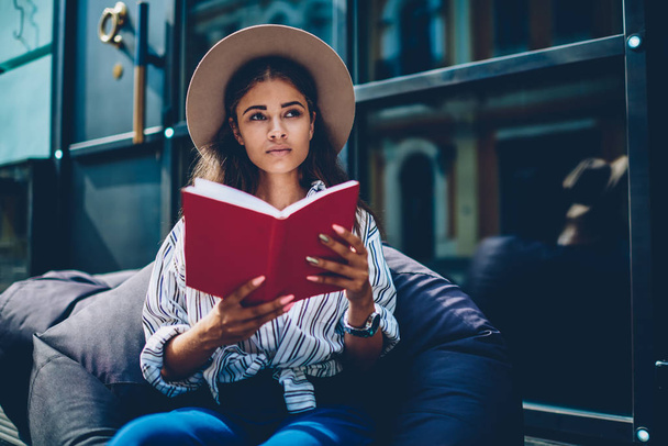 Thoughtful attractive talented hipster girl in hat on head thinking on plot of interesting novel read in red literature book.pondering student resting on pouf in coworking enjoying favourite bestseller - Photo, Image