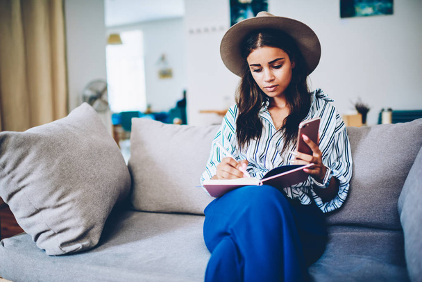 Stylish skilled designer driving sketch of trendy clothes in notepad sitting on soft sofa in cafe.Pensive businesswoman in hat writing down working plan in own coffee shop interior near copy space area - Foto, Imagen