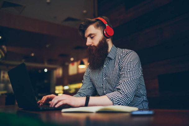 Professional bearded copywriter listening favourite music in headphones and typing text information on keyboard of modern laptop computer during distance job.Student in earphones working on coursework - Zdjęcie, obraz