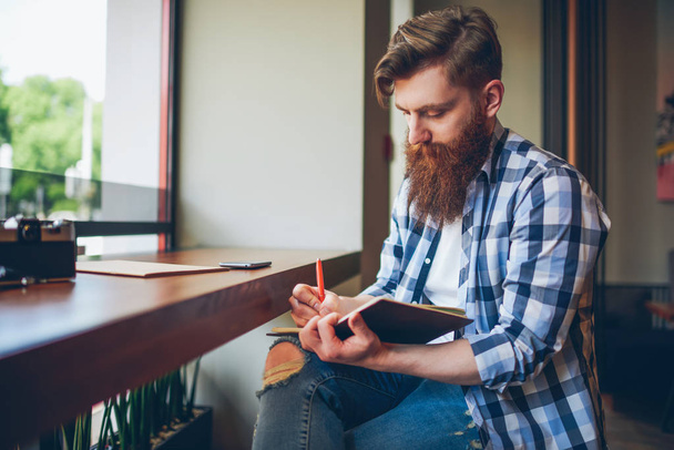 Talented young bearded man in casual shirt drawing sketch in notebook enjoying leisure time in coffee shop.Skilled student writing down homework in notepad sitting in coworking.Copy space area - Фото, изображение