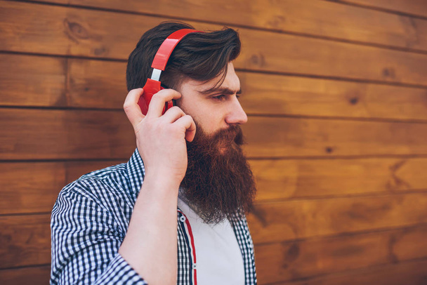 Vue latérale du jeune homme barbu élégant vêtu d'une chemise décontractée écoutant de la musique électronique préférée dans sa propre playlist via des écouteurs rouges modernes debout sur un fond promotionnel en bois
 - Photo, image
