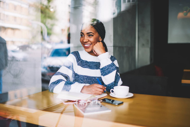 Dreamy black young woman thinking on new ideas for writing essay in notepad resting in stylish cafeteria interior.Smiling hipster girl working remotely on creating new successful project - Foto, afbeelding