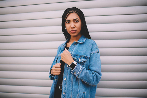 Half length portrait of pensive hipster girl dressed in casual denim apparel posing at camera standing on grey wall for text message.Black model with pigtails and smartwatch looking at camera - Foto, afbeelding