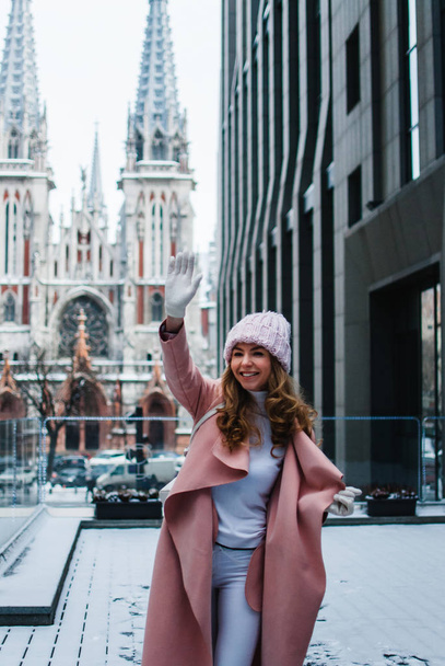 Dobrý den, z nového města! Krásná mladá žena v růžové kabátě mává a s úsměvem stoje venku při pohledu na fotoaparát. - Fotografie, Obrázek