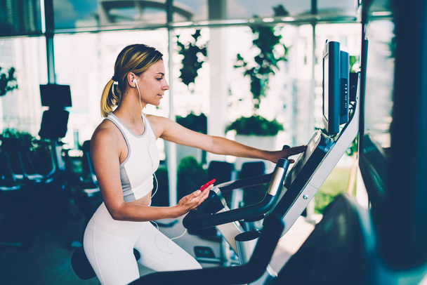 Attractive sportive woman choosing speed level on elliptical cross trainer while listening favourite music playlist on smartphone.Young fit female training in gym for supporting healthy lifestyle - Фото, изображение